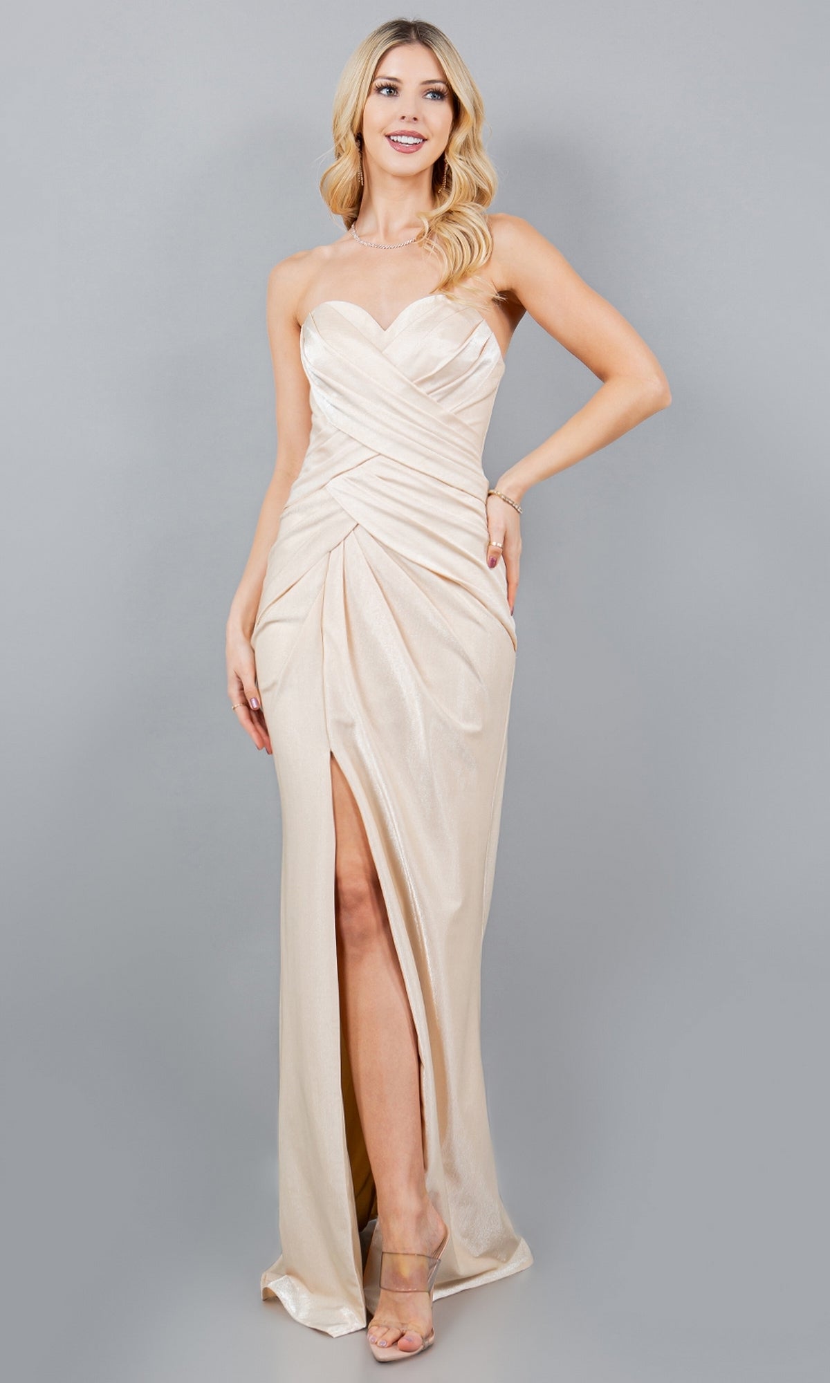 A woman wearing the Strapless Long Prom Gown: Cinderella Couture 8095J in light beige satin with a ruched bodice, sweetheart neckline, high slit, and clear-heeled sandals stands against a plain gray background.